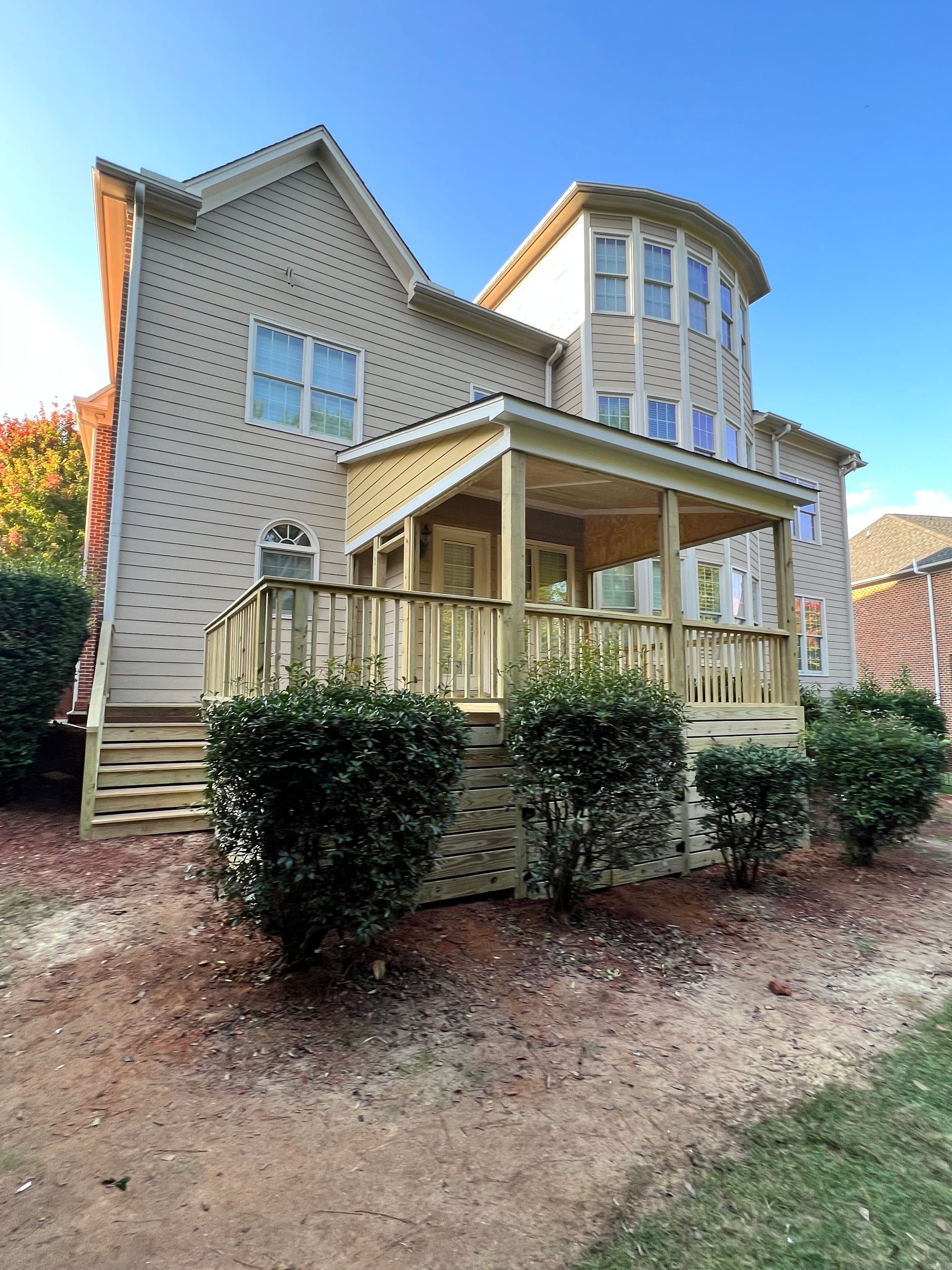 Back of a beige house with a wooden deck and porch. Shrubs in the foreground, blue sky.