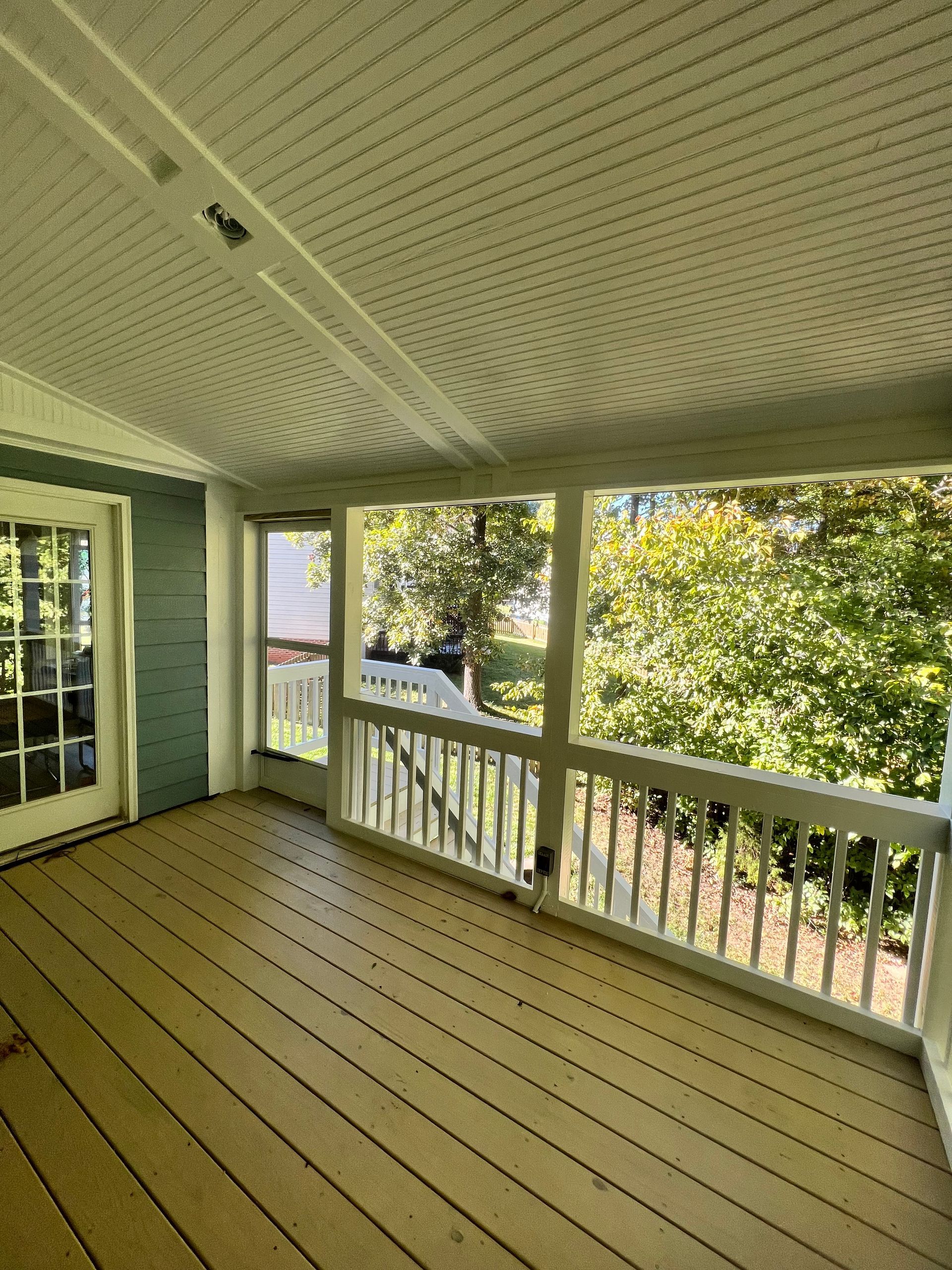 A screened-in porch with wood floor, white railing, and view of trees.