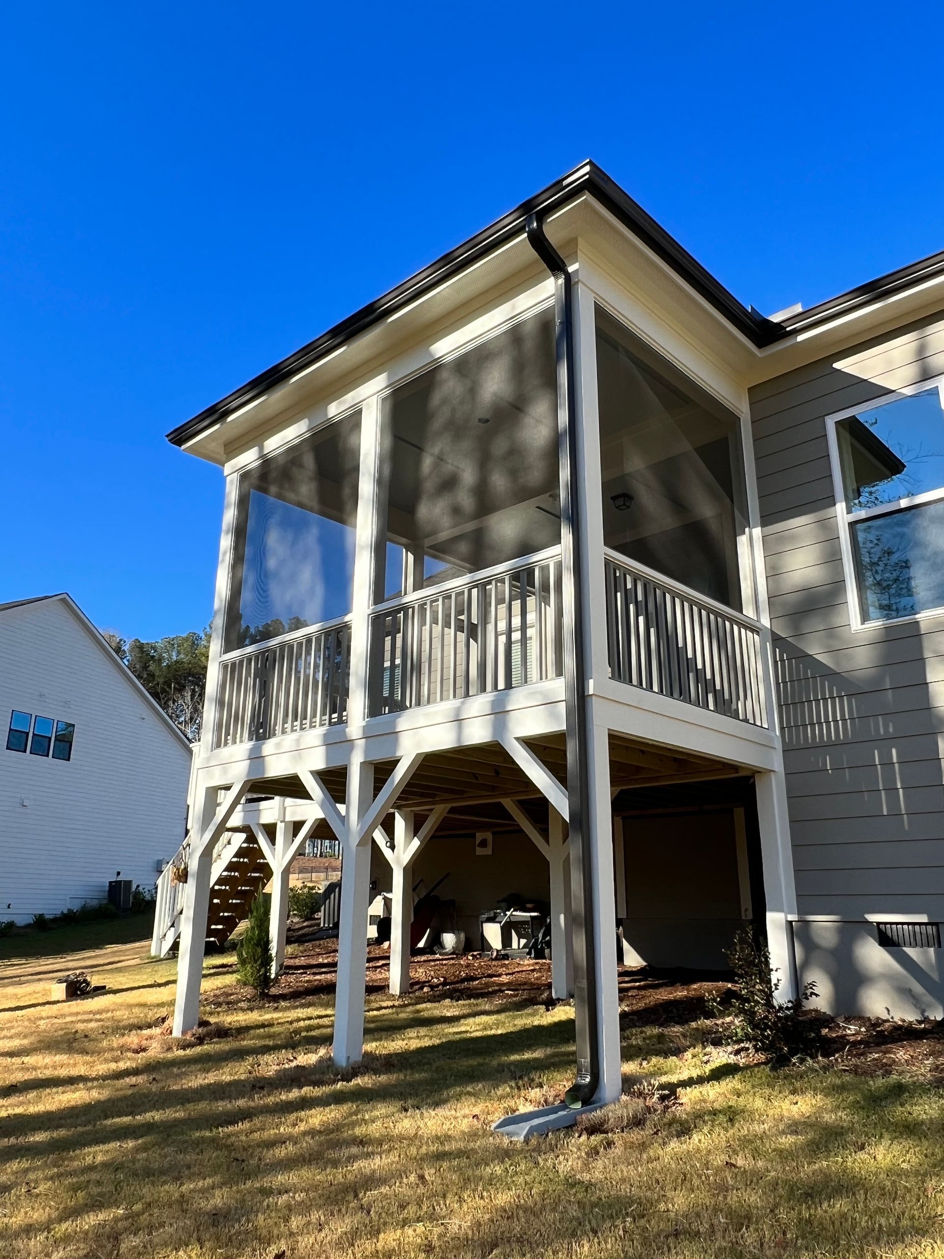 Screened-in porch on a house with white trim, wood railing, and gray siding; sunny day.