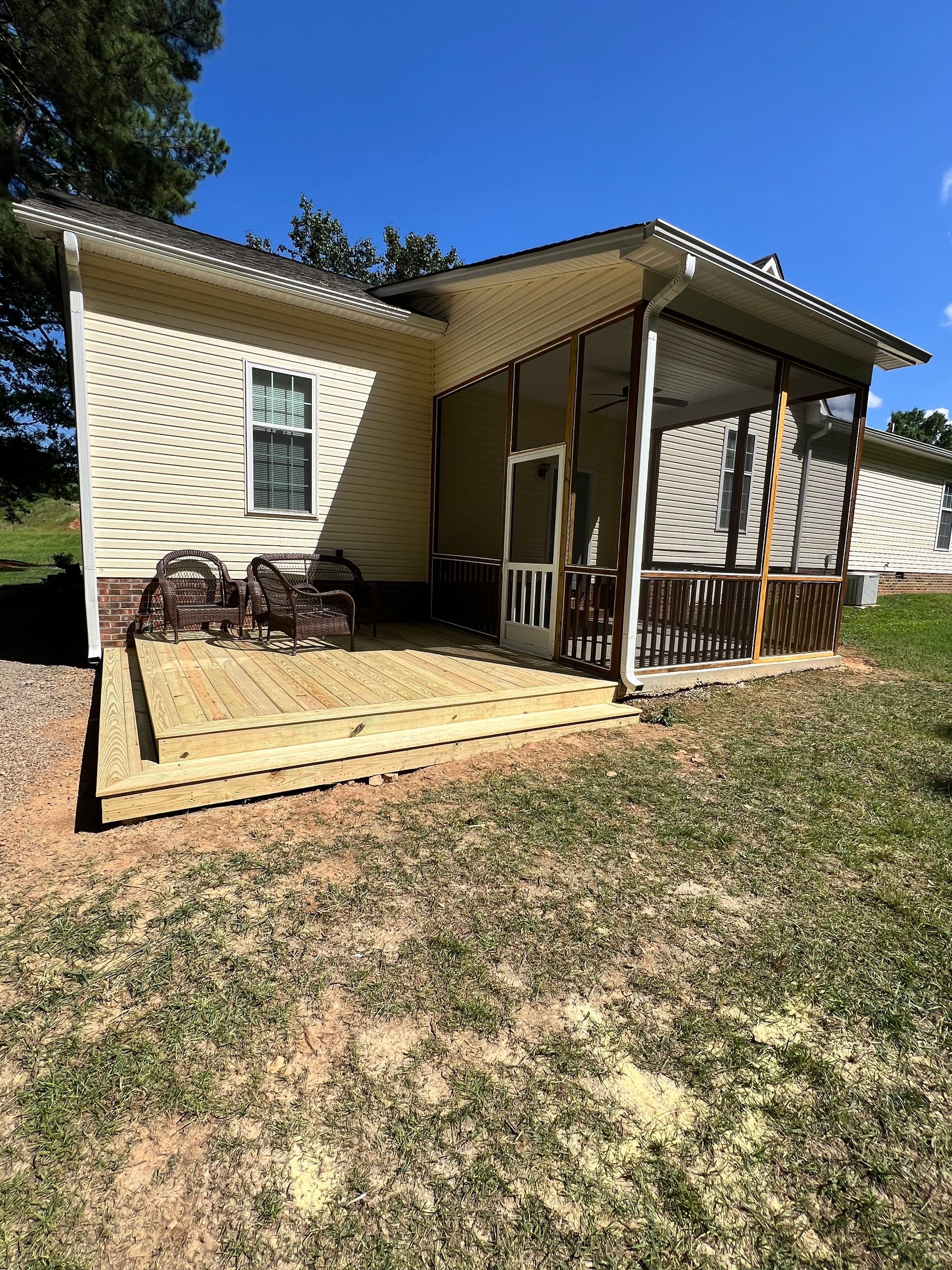A backyard with a wooden deck and screened porch attached to a yellow-sided house on a sunny day.