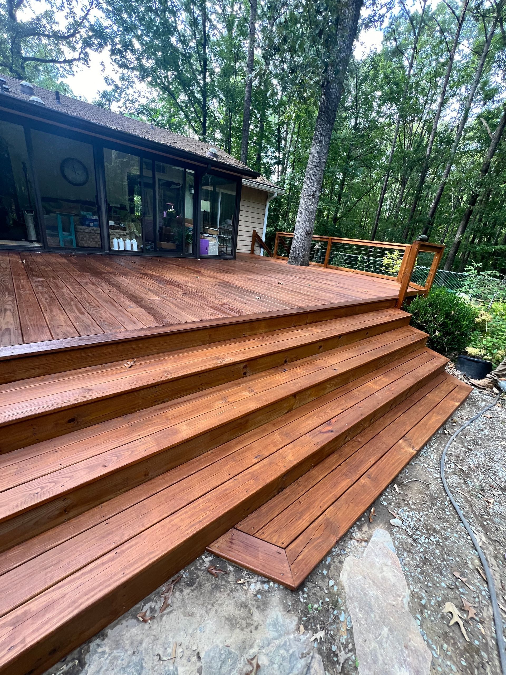 Newly stained wood deck with steps leading down to a gravel and stone area in a wooded setting.