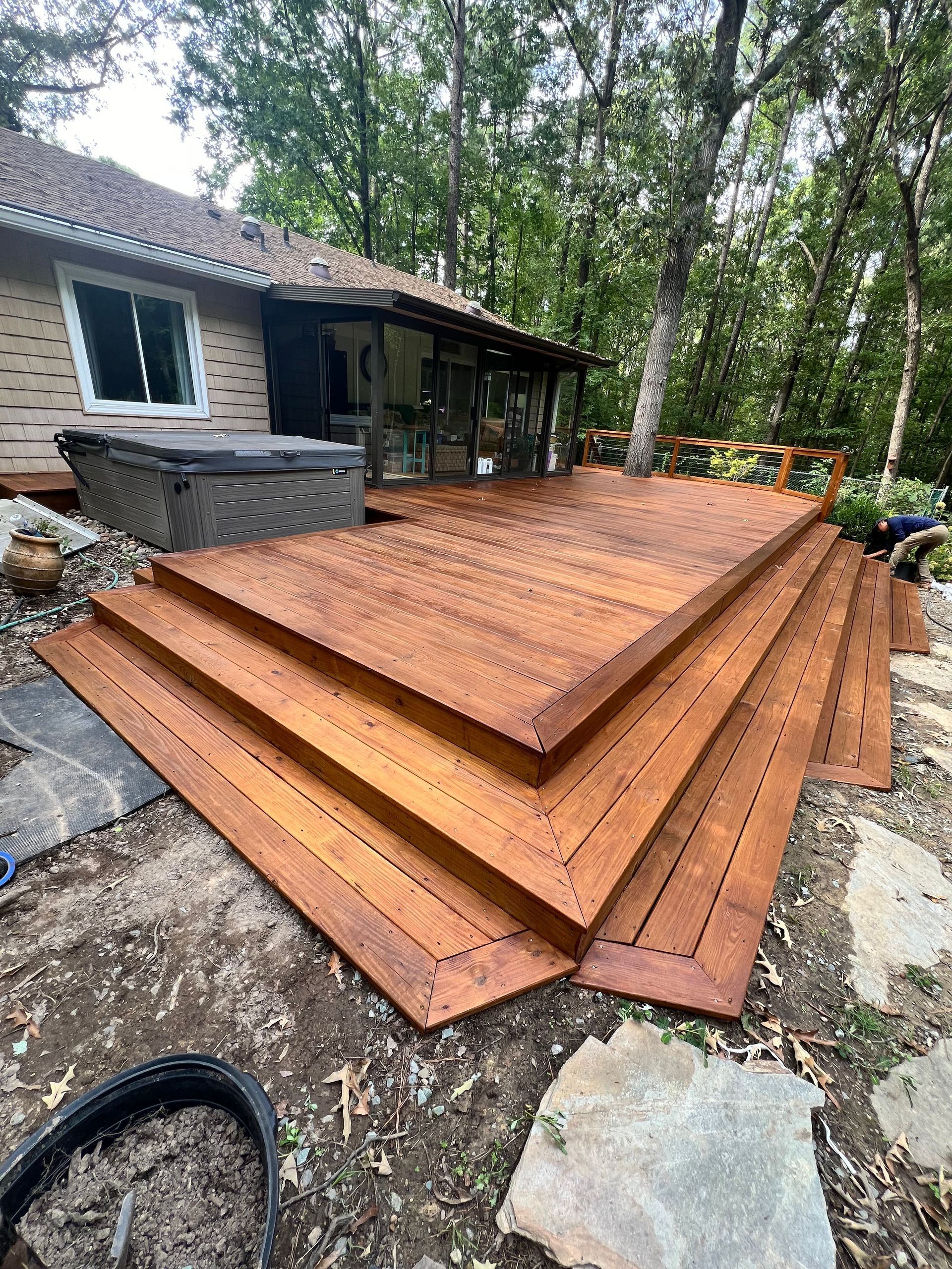 Wooden deck with steps next to a house and hot tub. Trees surround the backyard.