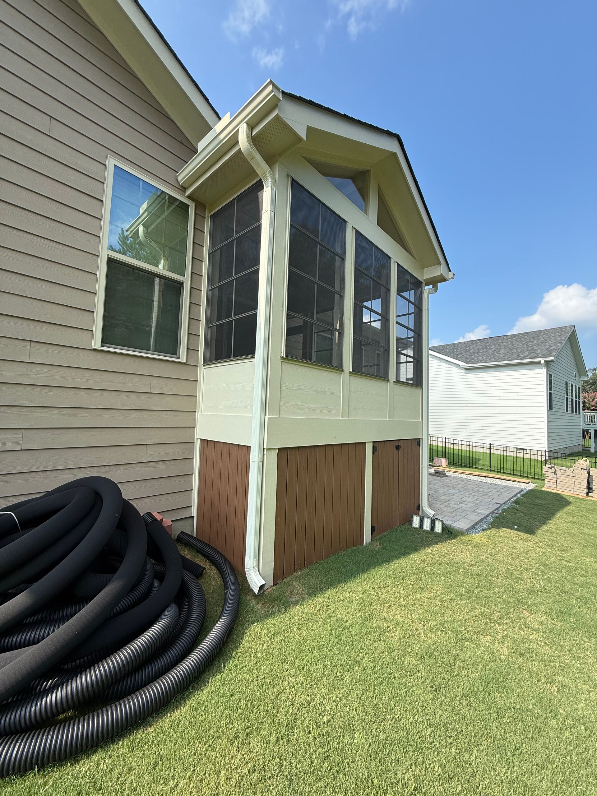 Screened porch with tan siding, brown base, and white trim next to a beige house, with coiled black tubing in the foreground.
