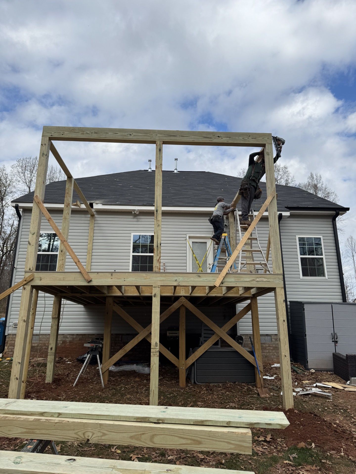Construction of a wooden deck. Workers install frame on the deck against a light colored house under a cloudy sky.