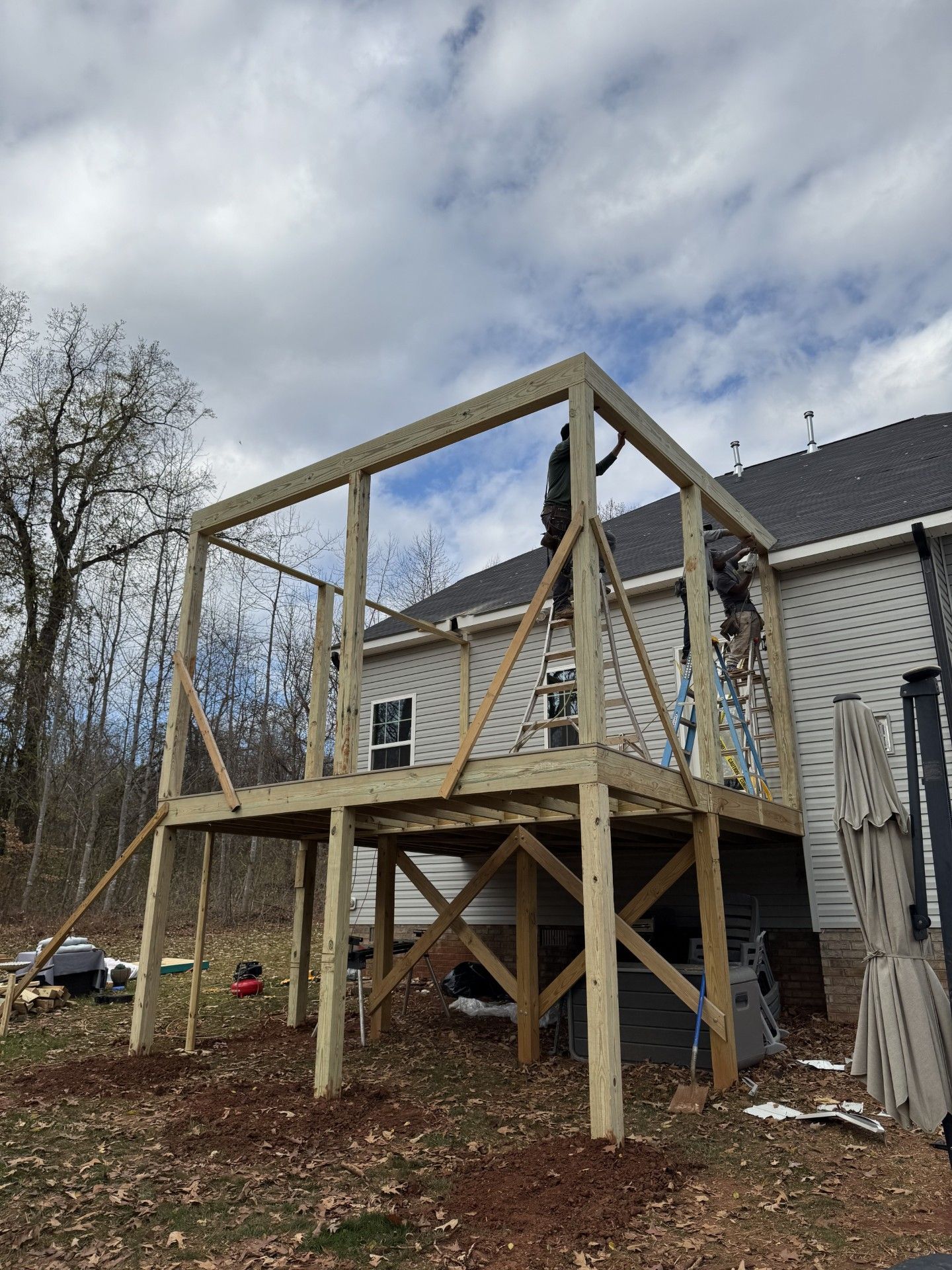 Wooden deck frame under construction with two workers, against a house and cloudy sky.