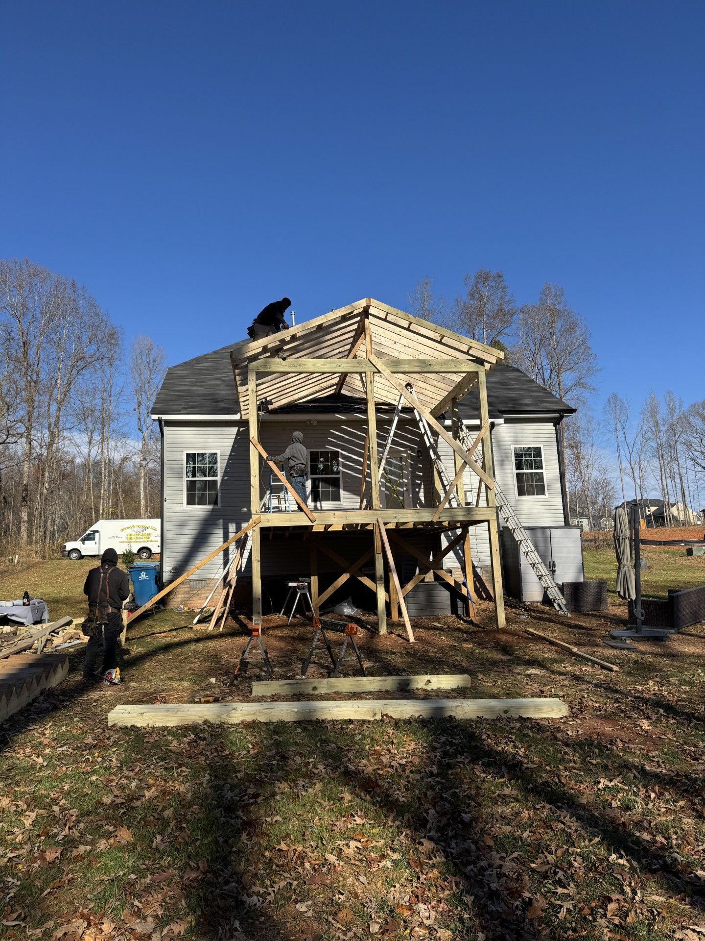 Deck being constructed on the back of a light gray house under a clear blue sky. Men are working on the structure.
