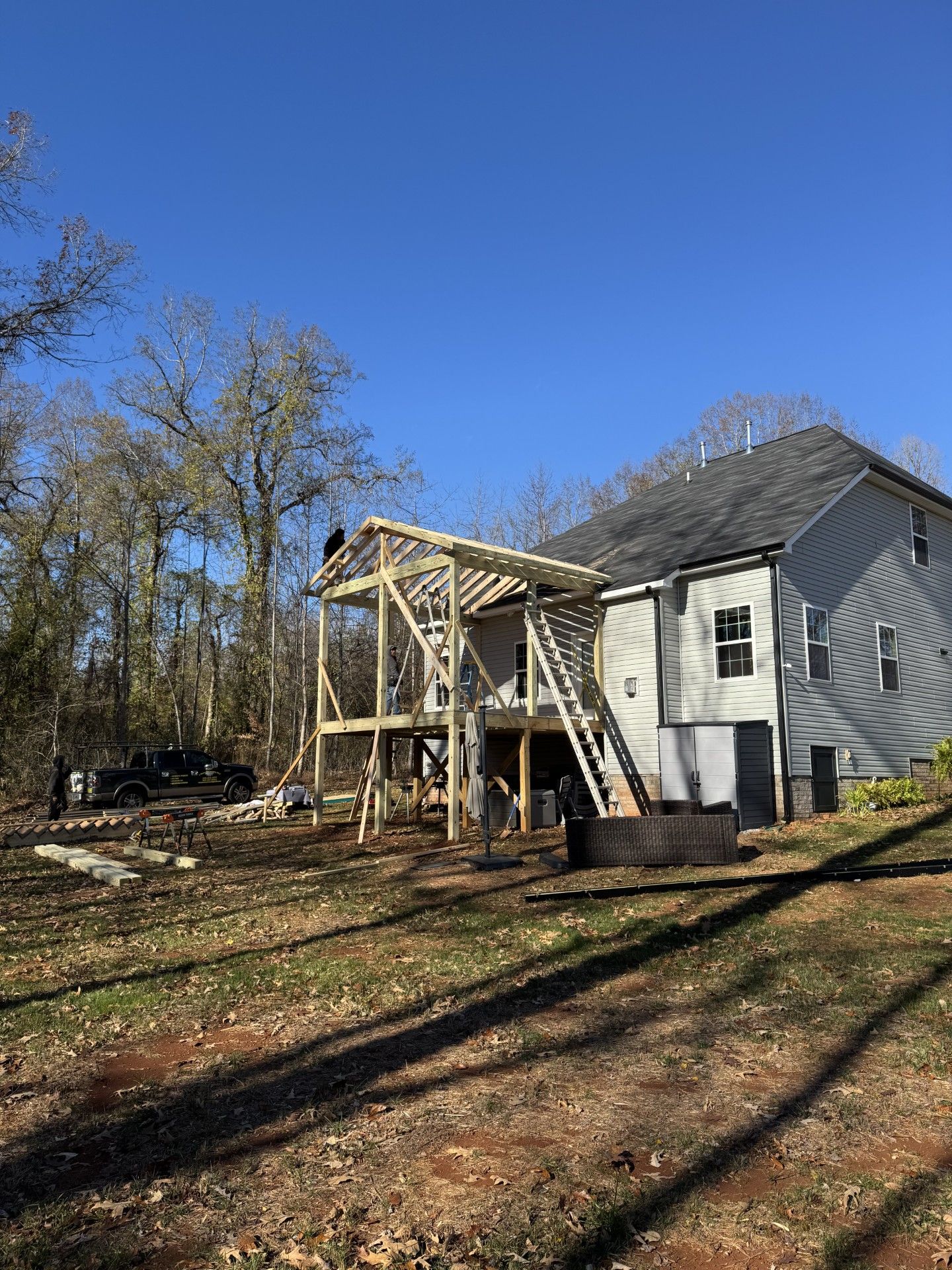 A wooden deck under construction is attached to a two story gray house. A sunny, outdoor setting.