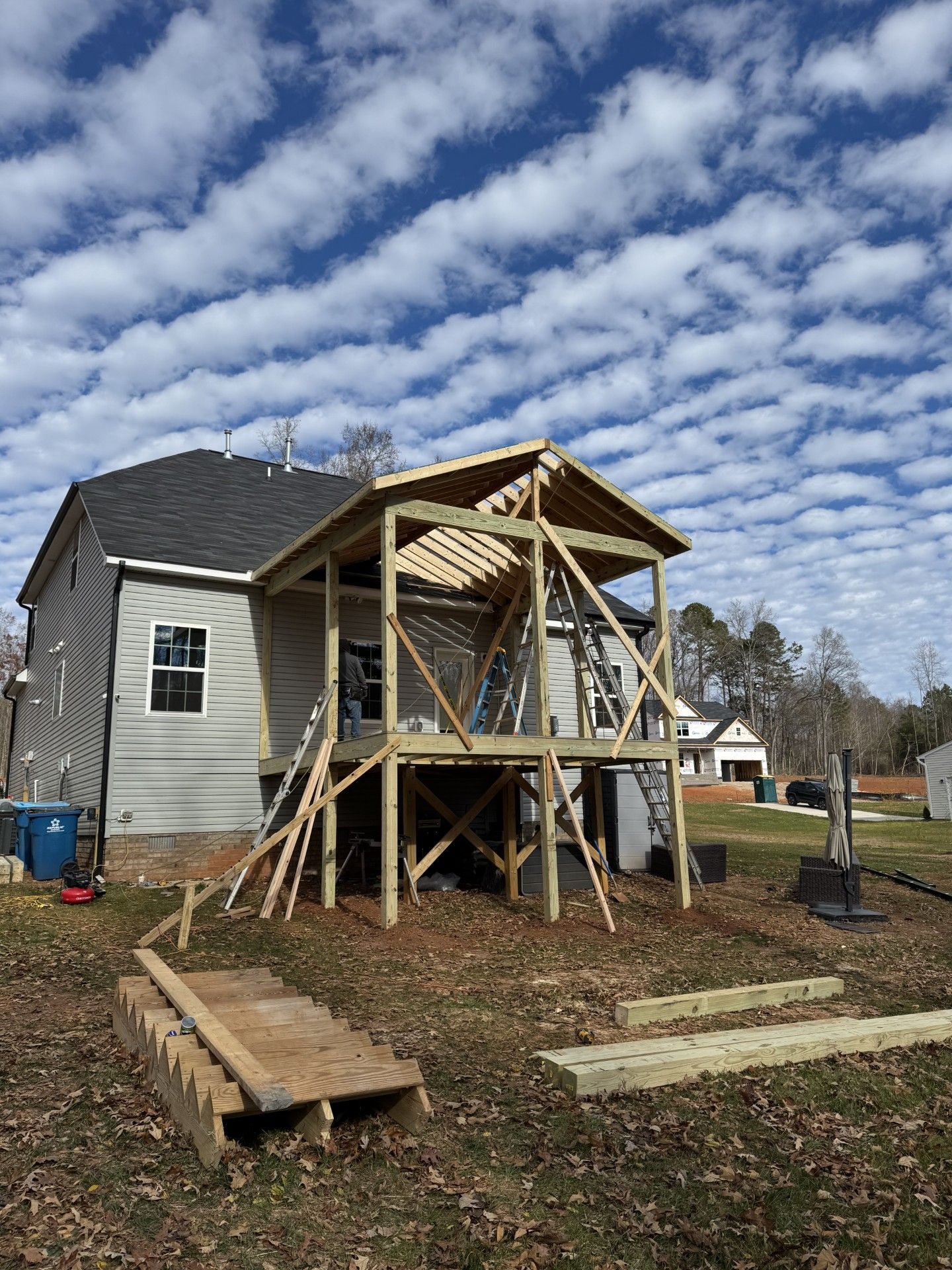 Wooden deck construction attached to a house with an unfinished roof, under a cloudy sky.