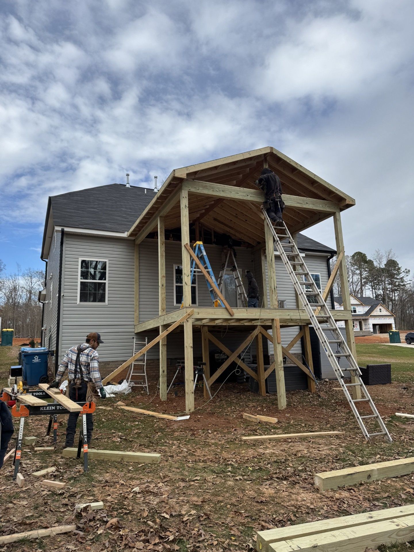 Construction workers building a covered wooden deck attached to a gray house under a cloudy sky.