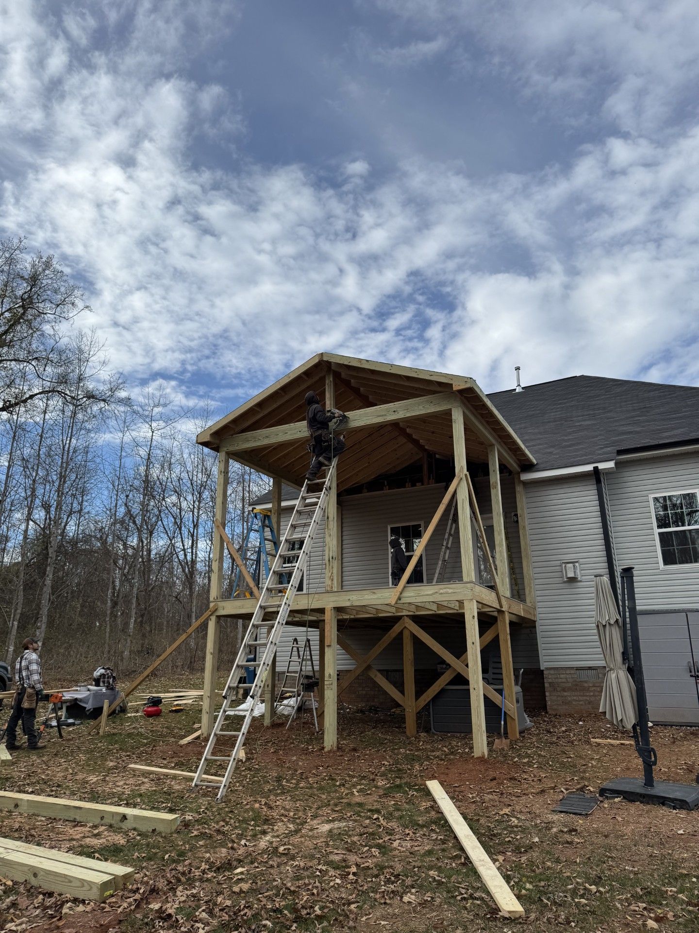 Construction of a wooden deck with a roof attached to a house; workers on a ladder, cloudy sky.