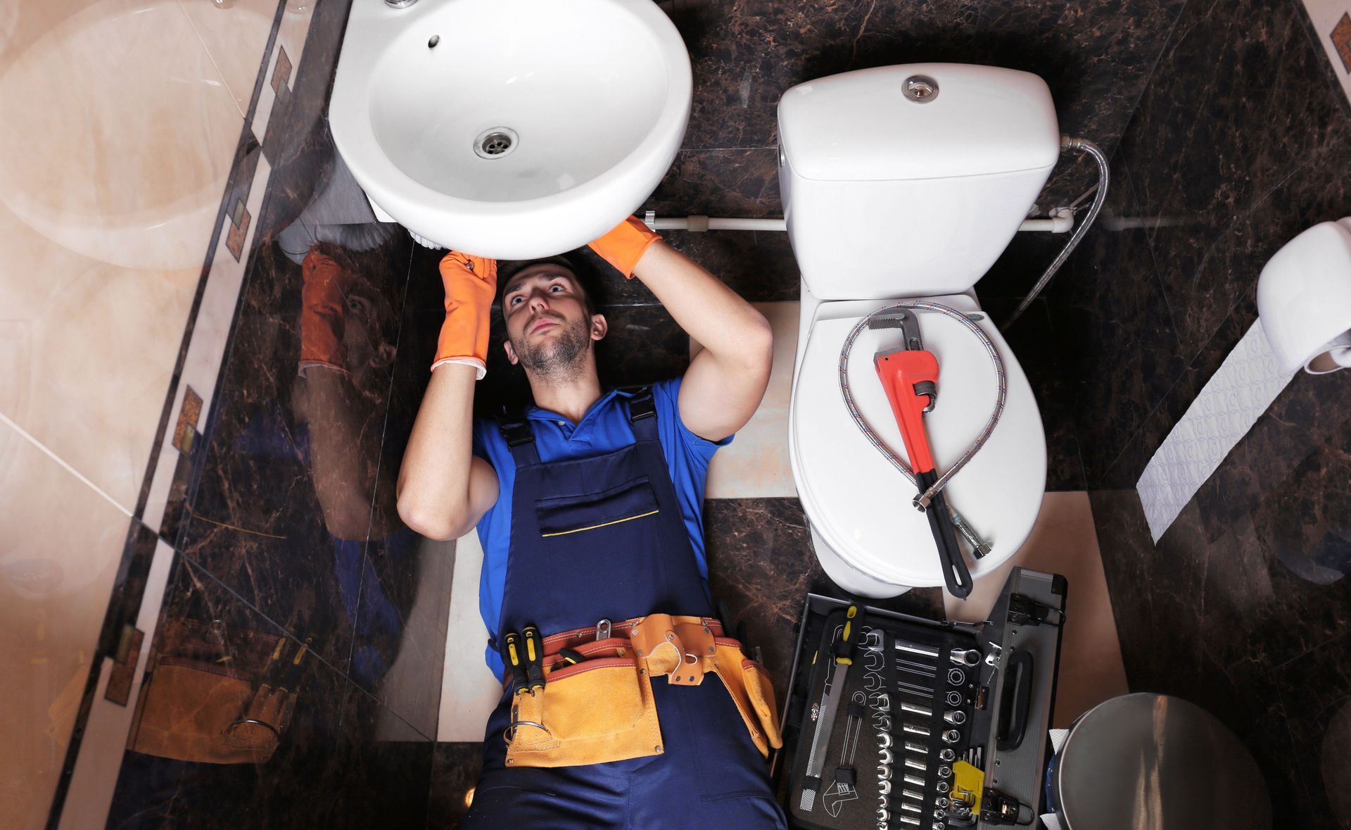 Plumber in blue overalls fixes sink in bathroom, with tools nearby, wearing orange gloves.