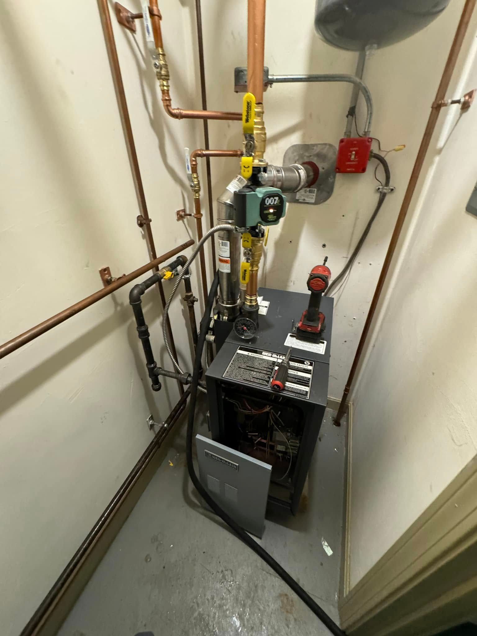 A boiler unit in a small room with copper pipes, wiring, and a power drill on top.