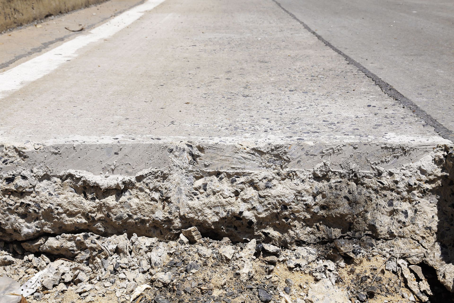 A close-up view of a damaged concrete slab on a roadway showing rough, crumbling layers and exposed aggregate.