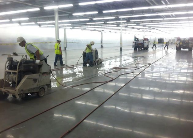 Construction workers in high-visibility vests operate concrete polishing machinery on a reflective warehouse floor.