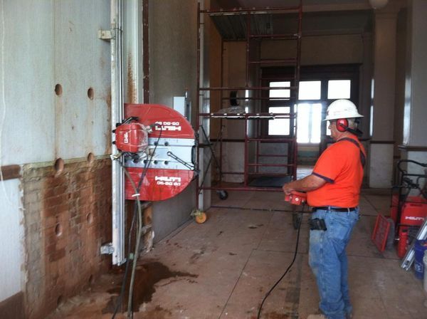 A worker in a high-visibility orange shirt uses a wall saw mounted on a metal track to cut through an interior wall.