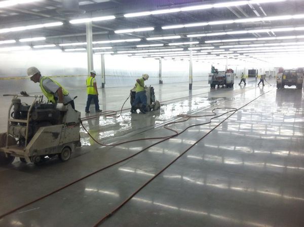Workers in high-visibility vests operate concrete floor polishing machines in a large, brightly lit industrial facility.