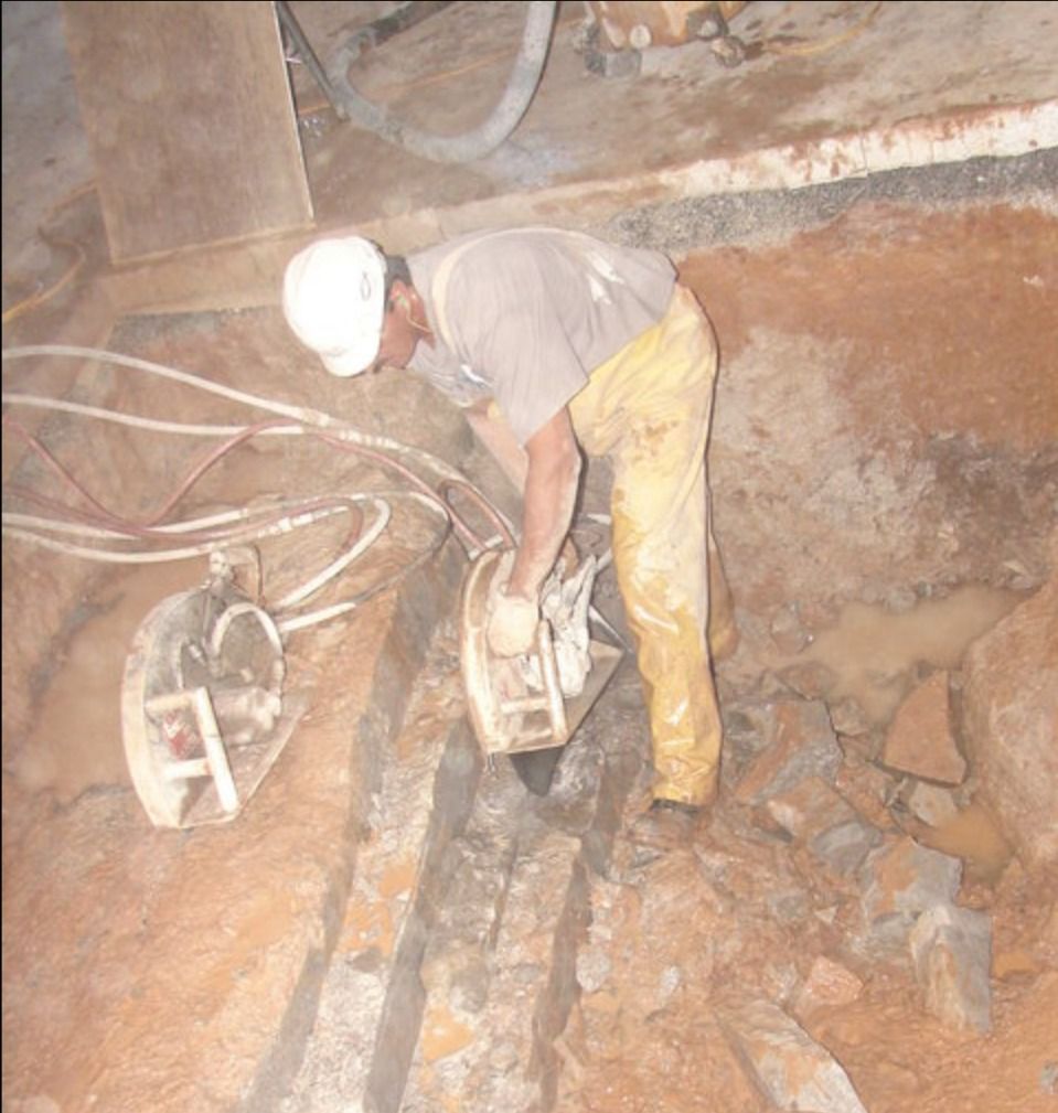 A worker wearing protective gear uses a large circular power saw to cut a deep trench into the dirt ground.