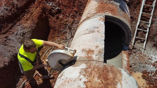 A worker in a high-visibility yellow shirt uses a concrete saw to cut a section out of a large pipe in a dirt trench.