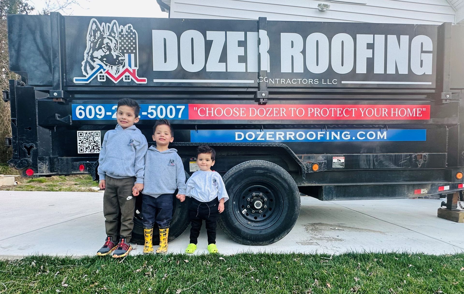Three young boys are standing in front of a dozer roofing trailer.