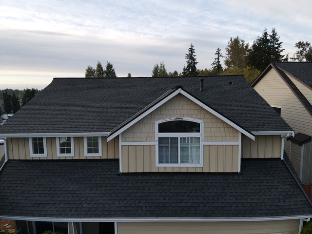 Black shingled roof on a two-story house with beige siding, white trim, and a window.