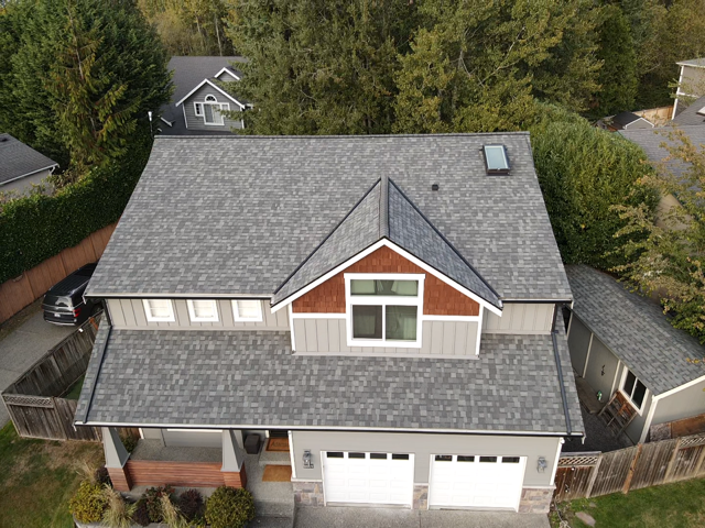 Gray-roofed house with two-car garage, brown trim, and a small skylight, surrounded by trees and a fence.