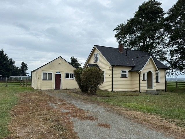 Yellow house and detached building with driveway on a grassy field under a cloudy sky.