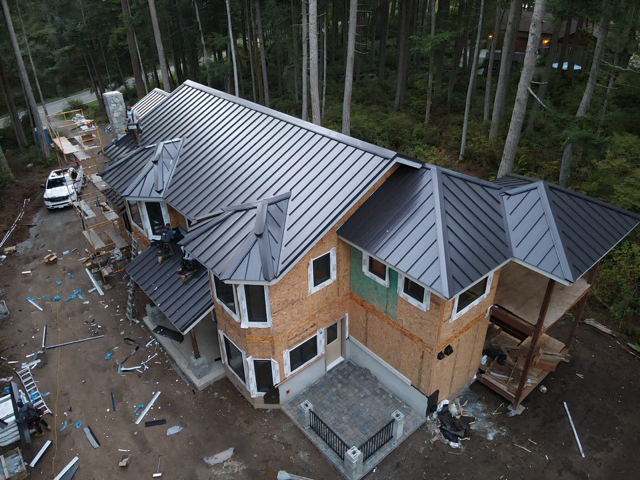 House under construction with gray metal roof, surrounded by trees. Workers on roof.