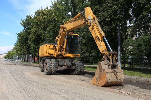 A yellow wheeled excavator stands on a gravel road alongside a line of trees.