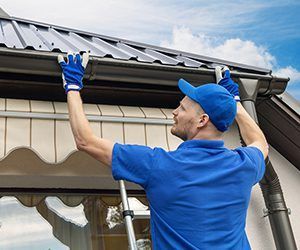 Worker in blue uniform cleaning a house gutter beside a roofline