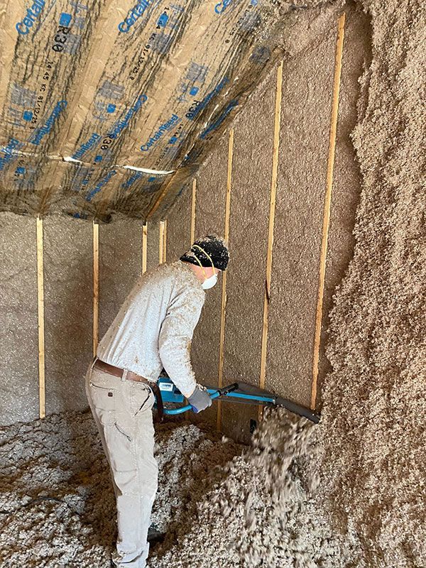 Person shoveling loose insulation inside an attic with exposed wooden rafters and hanging wires