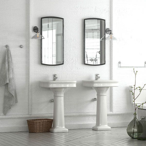 Bright white bathroom with twin pedestal sinks, mirrors, wall sconces, and a wicker basket.