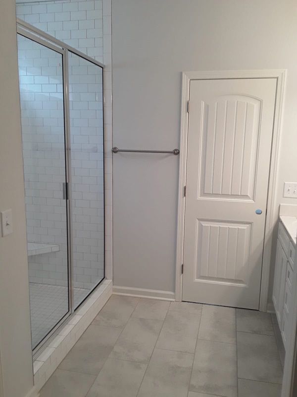 Bright bathroom with glass shower, gray tile floor, white door, and towel bar on a light gray wall