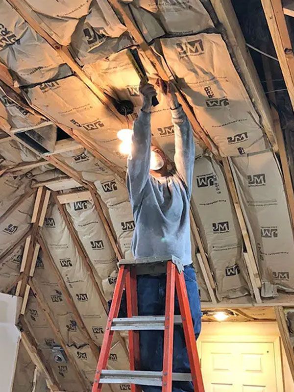 Worker on a ladder installing insulation in an attic with exposed beams and a lit bulb.
