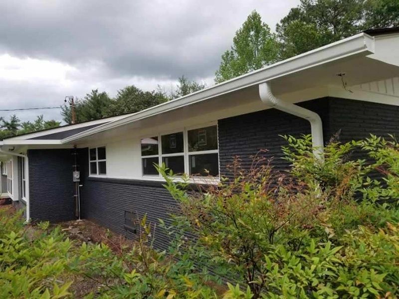 Dark brick house exterior with white trim, windows, overhanging roof, and shrubs in front.