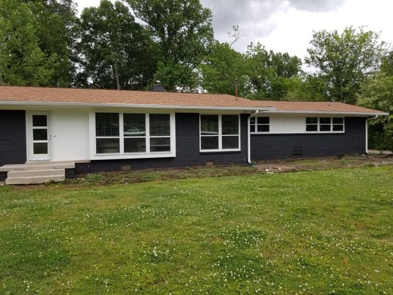 Single-story dark house with white trim, red roof, front steps, and a grassy yard under cloudy skies