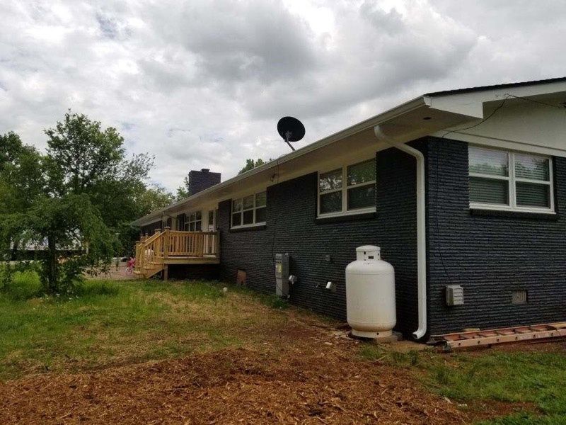 Dark gray house with a wooden deck, propane tank, and cloudy sky over a grassy yard