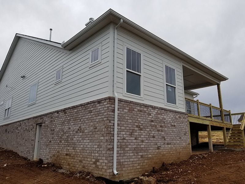 Side view of a raised house with white siding, brick foundation, and a wooden deck under a cloudy sky