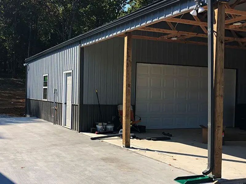 Metal workshop building with open bays, gray siding, and wooden posts beside a paved driveway