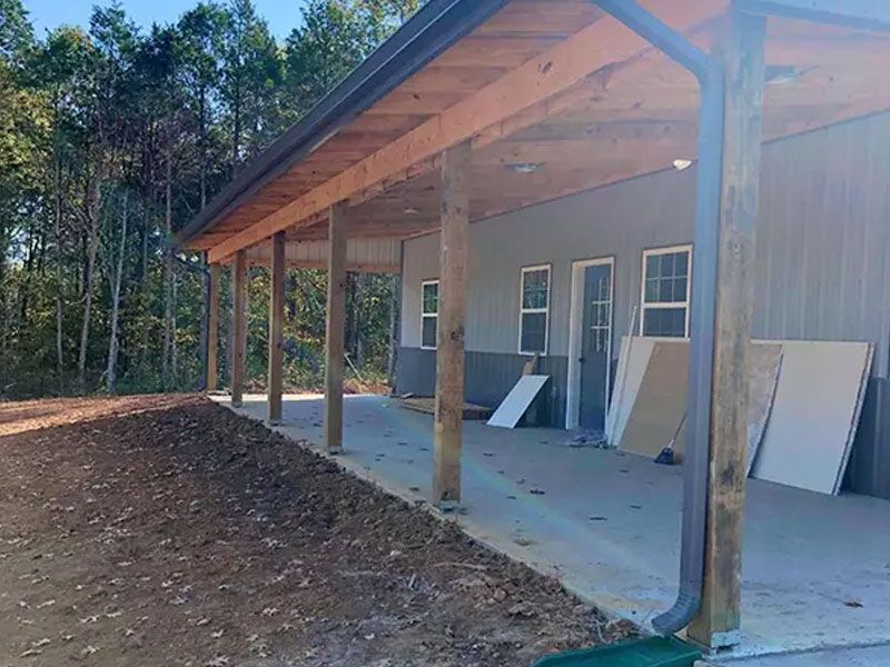 Covered porch of a house under construction, with lumber, panels, and a dirt yard beside trees.