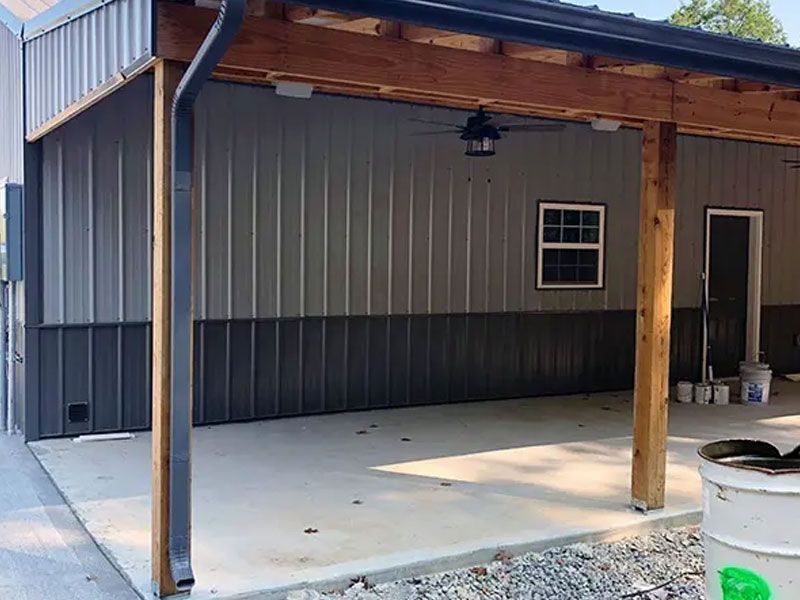 Covered concrete patio beside a gray metal building with wooden posts and a small window.