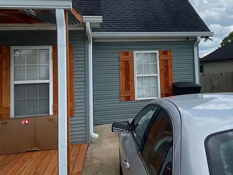 Porch with blue siding, white door and window, brown shutters, and a parked silver car in front