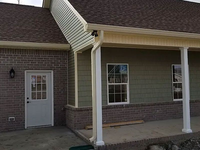Covered porch with white columns, tan siding, brick base, and a gray door on a house exterior.