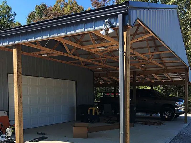 Open-sided carport with exposed wooden roof trusses, a white garage door, and parked vehicles inside