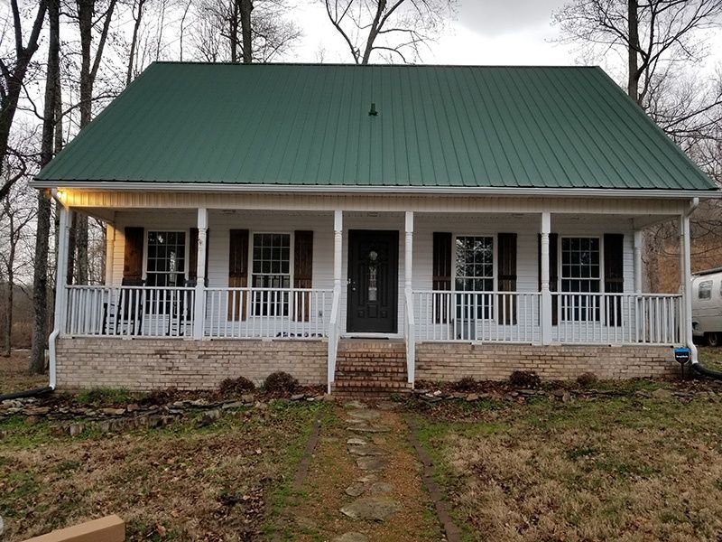 Small white house with a green metal roof and front porch in a wooded yard