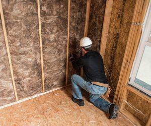 Worker kneeling inside a framed wall, installing insulation in an unfinished room with exposed wood studs.