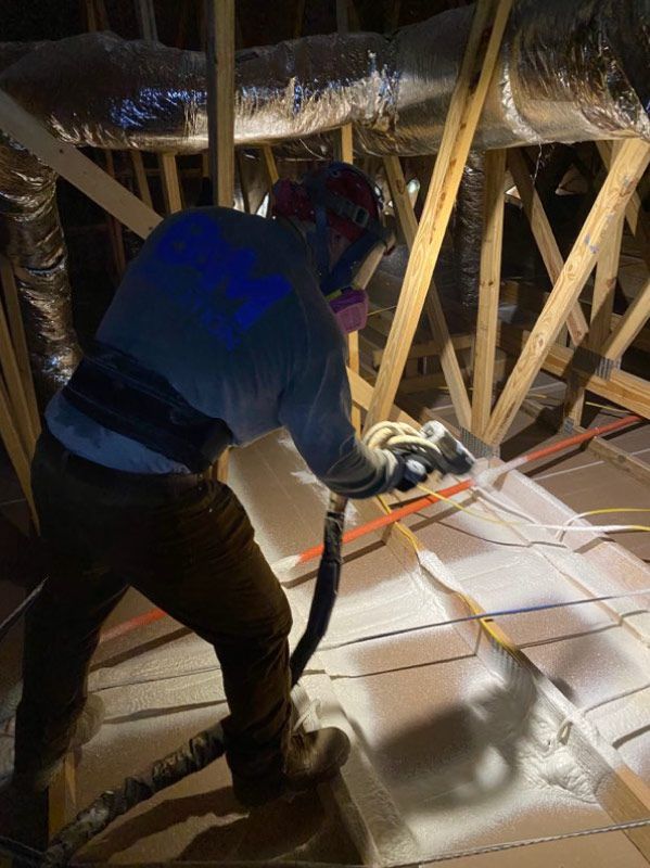Worker spraying insulation in an attic with exposed rafters and ductwork.