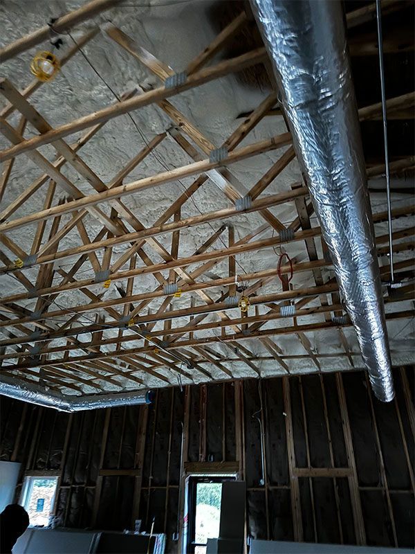 Exposed ceiling trusses and ductwork inside an unfinished room with open wall framing and windows.