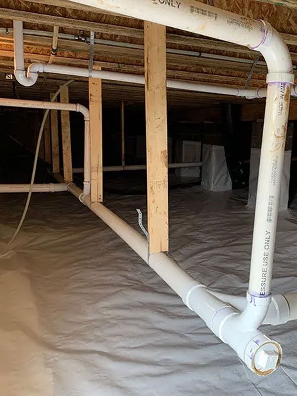 Crawlspace with exposed wooden joists and PVC plumbing pipes over a plastic vapor barrier.