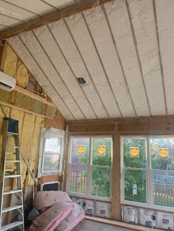 Unfinished room with exposed insulation, sloped ceiling, windows, ladder, and construction materials.