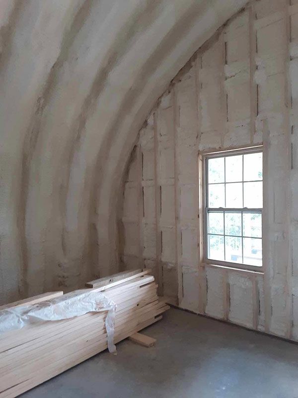 Unfinished attic room with exposed insulation, arched ceiling, stacked lumber, and a small window.